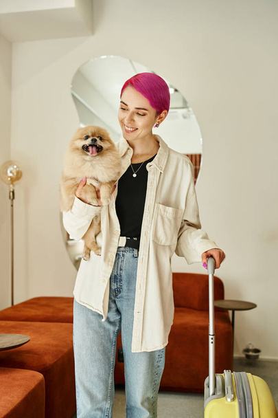 veselá žena drží legrační pomeranian spitz a cestovní taška v recepci psa hotelu - Fotografie, Obrázek