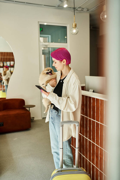 stijlvolle vrouw met pomeranian spitz en smartphone in de buurt van koffer en receptie in huisdier hotel - Foto, afbeelding