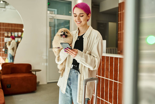 Küçük tüylü köpeği ve akıllı telefonu olan neşeli bir kadın resepsiyon masasının yanında ve köpek otelinde seyahat çantası taşıyor. - Fotoğraf, Görsel
