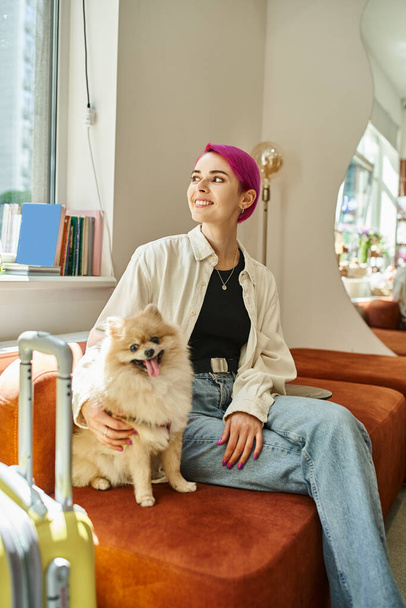smiley woman looking away while sitting with pomeranian spitz near suitcase in cozy pet hotel - Fotografie, Obrázek