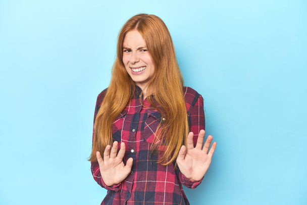 Redhead young woman on blue background rejecting someone showing a gesture of disgust. - Photo, Image