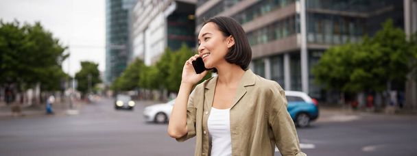 Muotokuva hymyilevä aasialainen nainen puhuu matkapuhelimella, kävelee kadulla lähellä vilkasta tietä ja puhuu ystävälle, nauraa. - Valokuva, kuva