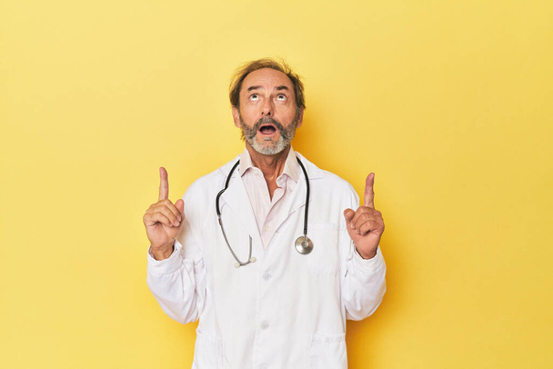Doktor se stetoskopem ve žlutém studiu ukazuje vzhůru nohama s otevřenými ústy. - Fotografie, Obrázek