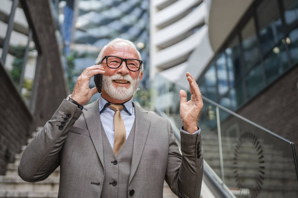 Портрет старшого чоловіка з бородатим бізнесменом у костюмі стоїть на відкритому повітрі перед сучасною будівельною корпорацією, використовуючи мобільний телефонний телефон, роблять розмову справжньою людиною - Фото, зображення