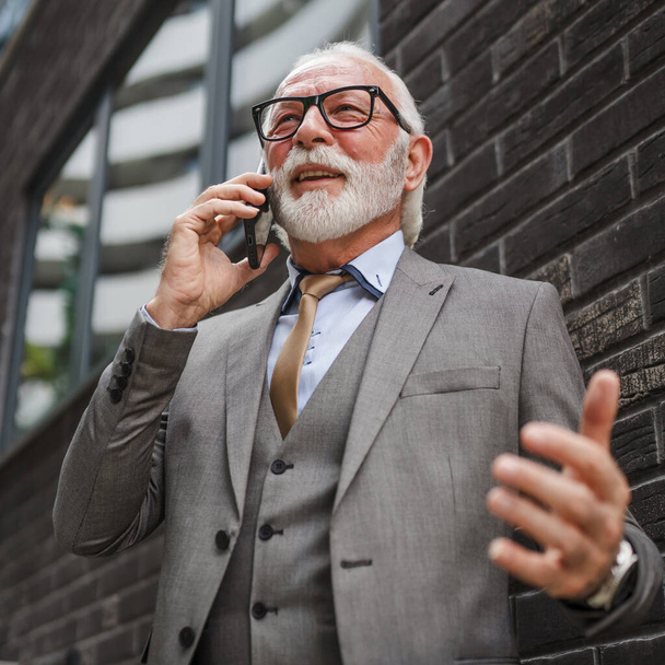 Πορτρέτο του ηλικιωμένου με γενειάδα επιχειρηματίας σε κοστούμι σταθεί υπαίθρια μπροστά από τη σύγχρονη εταιρεία κτίριο χρήση κινητό τηλέφωνο smartphone κάνουν μια κλήση μιλήσουμε πραγματικό πρόσωπο αντίγραφο χώρο - Φωτογραφία, εικόνα