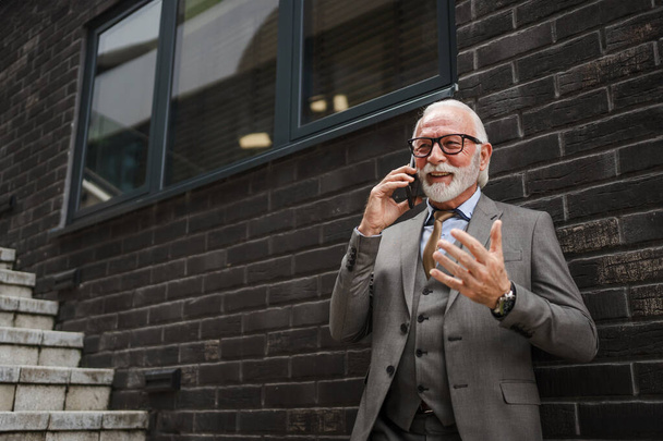Portrait of senior man with beard businessman in suit stand outdoor in front of modern building corporation use mobile phone smartphone make a call talk real person copy space - Photo, Image