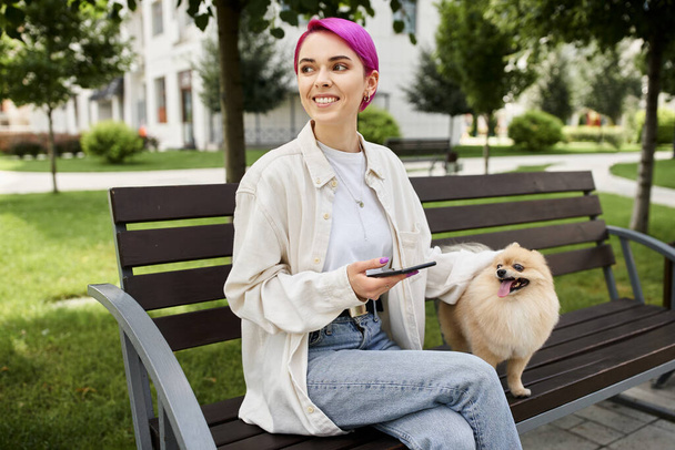 весёлая фиолетовая женщина со смартфоном, сидящая на скамейке в парке рядом с собакой - Фото, изображение