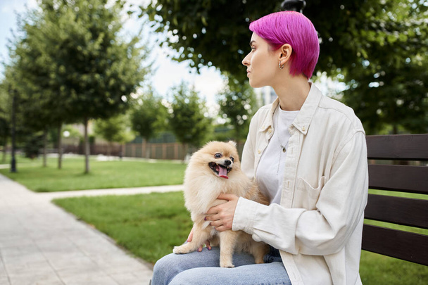 dreamy purple-haired woman with fluffy pomeranian spitz sitting on bench in park and looking away - Foto, Imagem