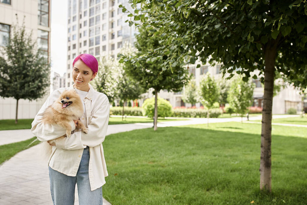 happy woman with trendy hairstyle walking on city street with pomeranian spitz in hands, enjoyment - Photo, Image