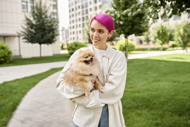 heureuse femme aux cheveux violets tenant un chien moelleux dans les mains et regardant loin dans la rue, promenade en ville - Photo, image