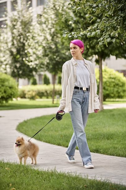 stylish woman looking away while walking in green city with pomeranian spitz on roulette leash - Фото, изображение