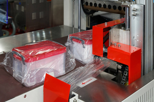 les boîtes de stockage en plastique sont enveloppées dans un film plastique transparent sur la bande transporteuse de la machine d'emballage dans la fabrication de récipients en plastique. - Photo, image