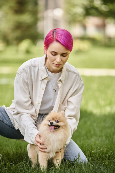 trendy purple-haired woman embracing cute pomeranian spitz on green grass in park, outdoor activity - Photo, Image