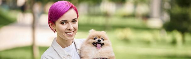 smiley and trendy woman with cute pomeranian spitz in hands looking at camera in park, banner - Photo, Image
