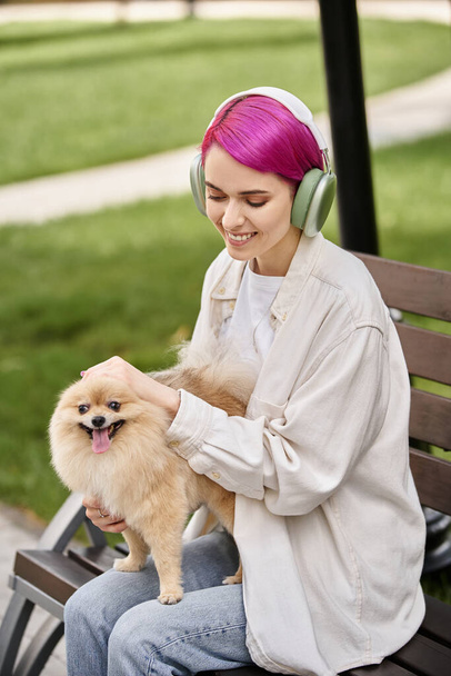 счастливая женщина слушает музыку в наушниках и гладит очаровательный поморский шпиц на скамейке в парке - Фото, изображение