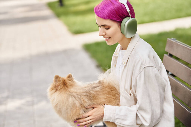 mujer alegre escuchando música en los auriculares y abrazando esponjoso spitz pomeranian en el banco en el parque - Foto, Imagen