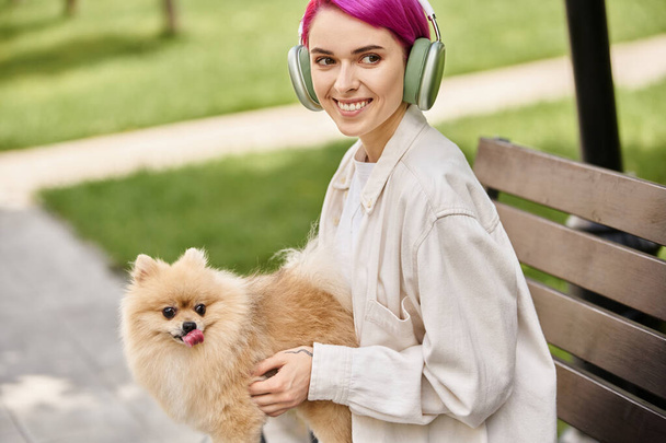 heureux femme aux cheveux violets assis dans le parc avec spitz poméranien et écouter de la musique dans les écouteurs - Photo, image