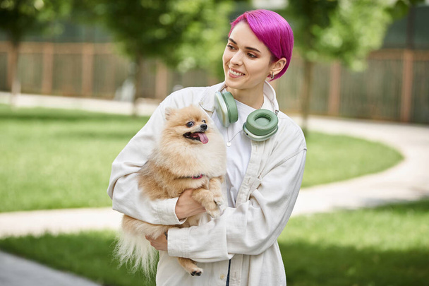 uśmiechnięta fioletowłosa kobieta ze słuchawkami spacerująca z uroczym psem czystej krwi w dłoniach na świeżym powietrzu - Zdjęcie, obraz