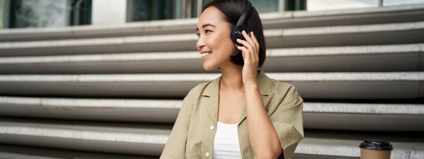 Nahaufnahme Porträt einer lächelnden asiatischen Frau hört Musik über Kopfhörer, dreht sich mit glücklichem Gesichtsausdruck um und ruht sich im Freien aus. - Foto, Bild