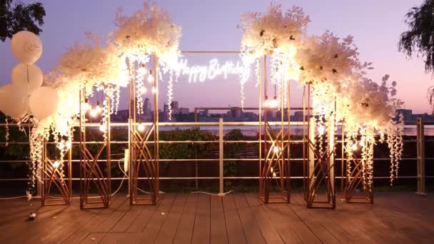 Iscrizione al neon Buon Compleanno su uno stand tra ghirlande di fiori bianchi con lampadine sullo sfondo del fiume sera Dnipro - Filmati, video