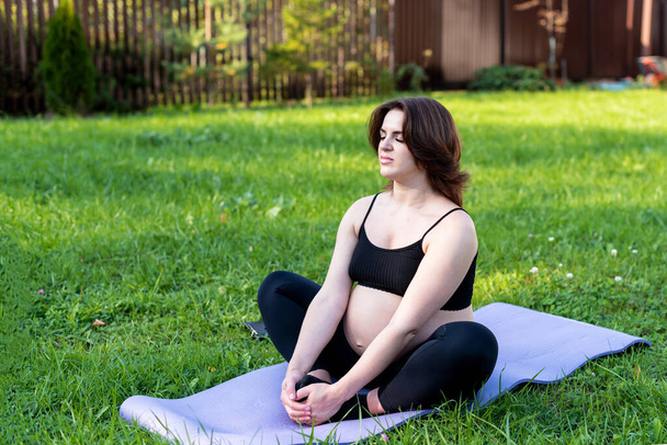 Hamile bir kadın yoga yapıyor ve bahçede çimenler üzerinde meditasyon yapıyor, temiz havayla derin bir nefes alıyor yoga nilüferleri rahat poz veriyor. Yoga Anneliği, Akıl Sağlığı ve Hamile Konsepti. - Fotoğraf, Görsel