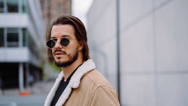 Portrét mladého atraktivního muže v neformálním oblečení, nošení slunečních brýlí a pózování na ulici před moderními budovami - Fotografie, Obrázek