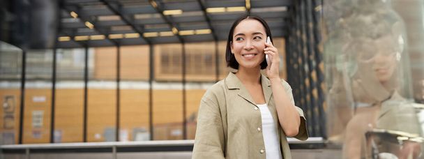 Gelukkige Aziatische vrouw aan het praten op mobiele telefoon, staande op de bushalte in de stad. Buitenschot. - Foto, afbeelding