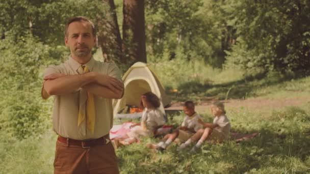 Portrait ralenti moyen d'un chef scout caucasien d'âge moyen confiant posant pour un appareil photo les mains repliées pendant l'été avec des enfants dans un camp forestier - Séquence, vidéo