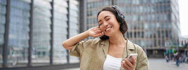 Šťastná mladá žena tančí na ulicích a poslouchá hudbu ve sluchátkách, drží smartphone. - Fotografie, Obrázek