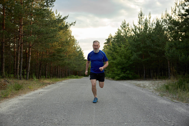 Hombre mayor corriendo fuera entre la carretera del campo. Estilo de vida saludable y actividad física para el bienestar en la jubilación - Foto, imagen