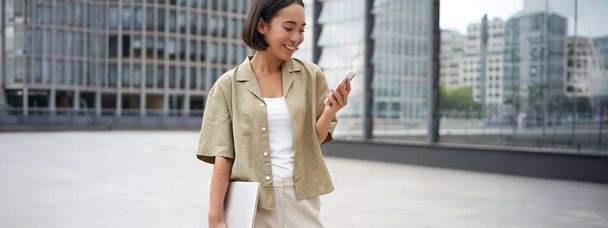 Junge asiatische Frau mit Laptop, die auf der Straße läuft und SMS schreibt, lächelt, während sie auf ihr Smartphone schaut. - Foto, Bild