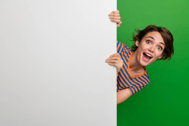 Φωτογραφία πορτρέτο της ελκυστική νεαρή γυναίκα κρύβονται πίσω από λευκό πανό τοίχου φορούν μοντέρνα ριγέ ρούχα που απομονώνονται σε πράσινο χρώμα φόντο. - Φωτογραφία, εικόνα