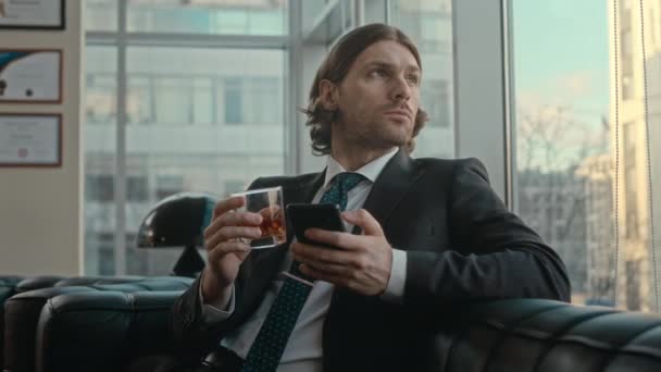 Vita fino di fiducioso giovane uomo d'affari caucasico in abito elegante utilizzando smartphone e bere whisky seduto sul divano in pelle nel suo ufficio di lusso - Filmati, video