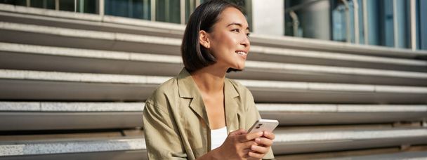 Ihmiset ja teknologia. Hymyilevä kaunis aasialainen nainen istuu portaissa kaupungissa, pitäen matkapuhelin. Tyttö älypuhelin lepää ulkona. - Valokuva, kuva
