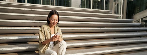 Kaunis aasialainen tyttö istuu portaissa rakennuksen ulkopuolella, matkapuhelimella, katselee älypuhelimen sovellusta ja hymyilee. - Valokuva, kuva
