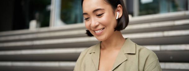 Portret van lachend aziatisch meisje luistert muziek, podast in draadloze oortelefoons, gebruik koptelefoon buiten, zittend op straat. - Foto, afbeelding