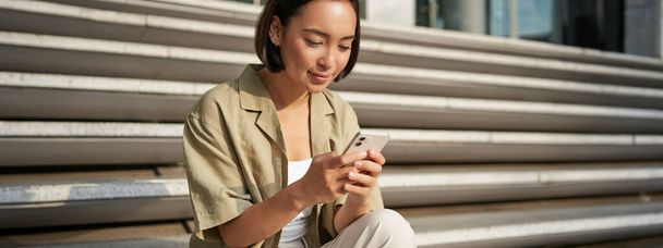 Menschen und Technologie. Lächelnde schöne asiatische Frau, die auf einer Treppe in der Stadt sitzt und ihr Handy in der Hand hält. Mädchen mit Smartphone ruht draußen. - Foto, Bild