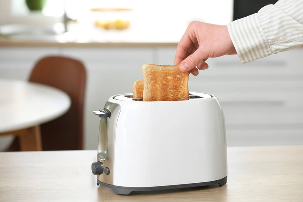 Mann nimmt knuspriges Brotstück aus modernem Toaster auf Tisch in Küche - Foto, Bild