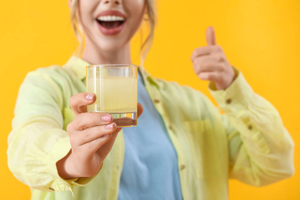Mulher jovem com comprimido dissolvido em vidro de água mostrando polegar para cima no fundo amarelo, close-up - Foto, Imagem