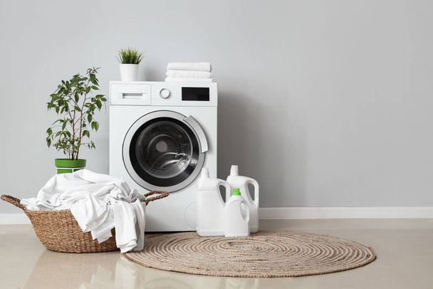 Máquina de lavar, garrafas de detergente e cesta com roupa suja perto da parede cinza - Foto, Imagem