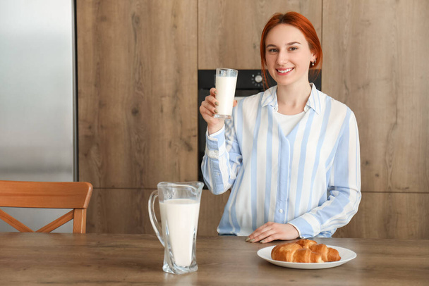 Молода руда жінка зі склянкою молока і круасан на кухні - Фото, зображення