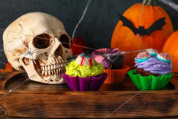 Board with delicious Halloween cupcakes and skull on wooden background, closeup - Photo, Image