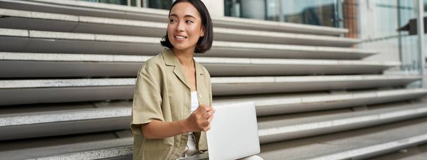 Портрет молодой женщины-студентки, азиатки с ноутбуком. Назначить улыбающейся девушке, цифровой кочевник работает над своим проектом удаленно, сидя на улице. - Фото, изображение