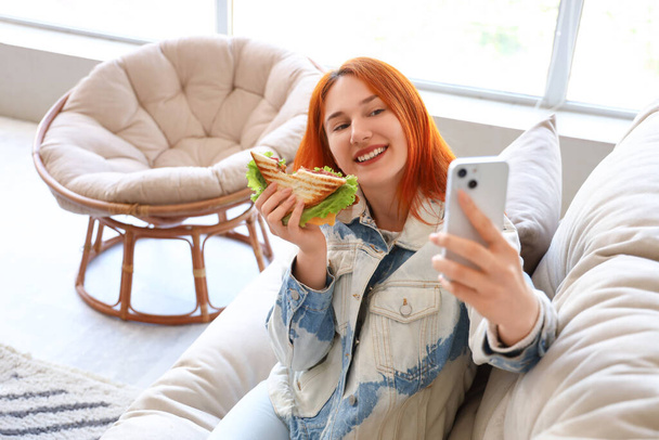 Joven pelirroja con sabroso sándwich tomando selfie en casa - Foto, imagen