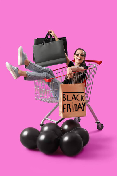 Krásná mladá žena s taškami a balónky sedí v nákupním košíku na fialovém pozadí. Černý pátek prodej - Fotografie, Obrázek