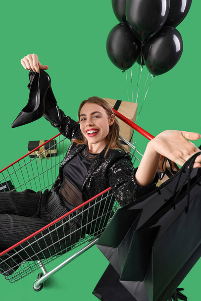 Young woman with bags and heels in shopping cart on green background. Black Friday sale - 写真・画像
