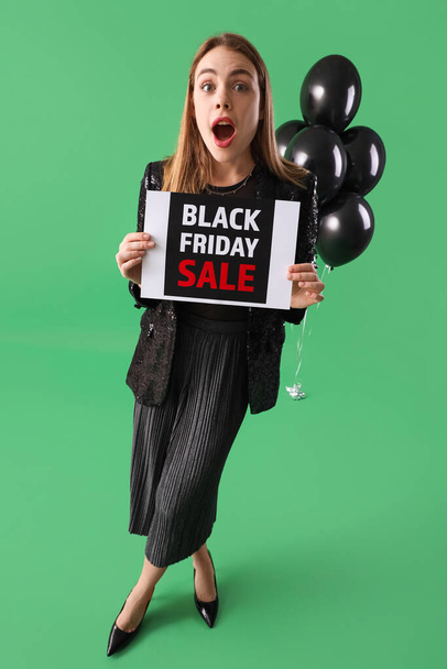 Surprised young woman with card and balloons on green background. Black Friday sale - 写真・画像