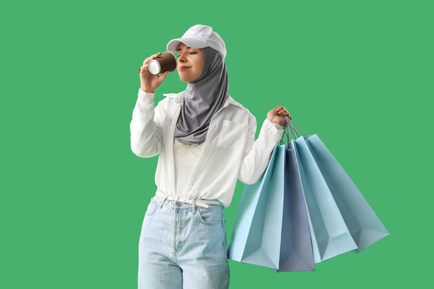 Młoda muzułmanka z torbami na zakupy i filiżanką kawy na zielonym tle - Zdjęcie, obraz