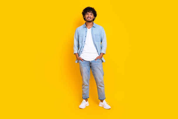 Full size photo of satisfied cool man with curly hairdo dressed denim shirt jeans hold arms in pockets isolated on yellow color background. - Photo, Image