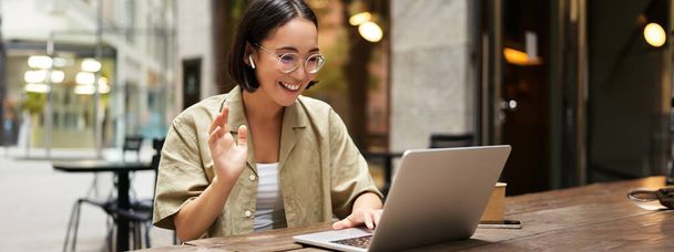 Çevrimiçi toplantı. Genç şehirli kız, Asyalı kadın dizüstü bilgisayarla konuşuyor, görüntülü konuşuyor, el kol hareketi yapıyor, açık bir kafede oturuyor ve uzaktan çalışıyor.. - Fotoğraf, Görsel